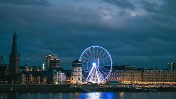 Illuminated Ferris wheel near riverside buildings at night.