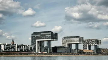 Modern buildings by a riverside, cloudy sky.
