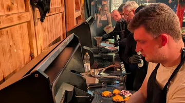 People cooking at grill inside wooden cabin.