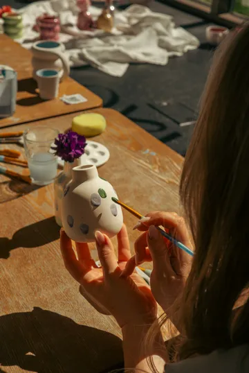 Woman painting a vase at a table.