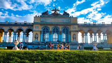 Menschen gehen nahe historischem Gebäude, blauer Himmel.