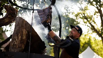 Chef grilling meat over open fire outdoors.
