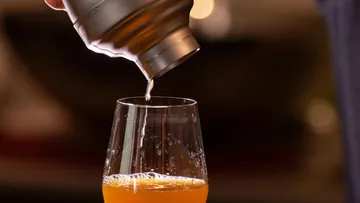 Shaker gießt Getränk in Glas, verschwommener Hintergrund.