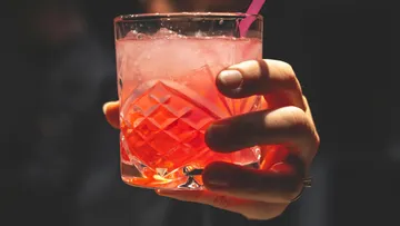Hand holding glass with pink drink, straw inserted.