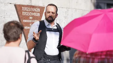 Mann im Anzug spricht draußen unter pinkem Regenschirm.