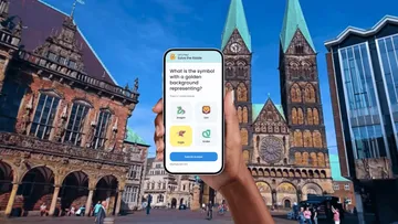 Hand hält Handy mit Quiz-App auf Stadtplatz.