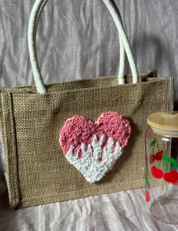 Gewebte Tasche mit Herzdesign, Glasgefäß daneben.