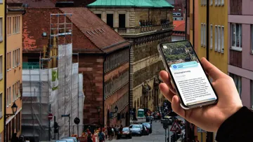 Hand holds smartphone, city street in background.