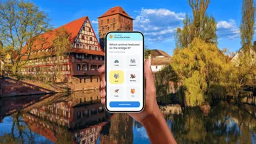 Hand hält Telefon vor Fachwerkhaus.