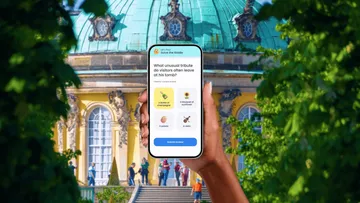 Hand hält Handy mit Quiz vor historischem Gebäude.