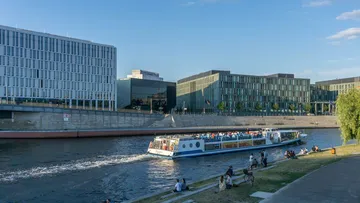 Tourboot fährt auf Fluss, moderne Gebäude dahinter.