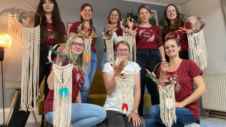 Frauen präsentieren stolz handgefertigte Makramee-Kunstwerke drinnen.