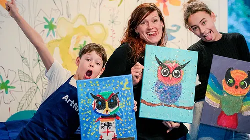 Three people holding colorful owl paintings indoors.