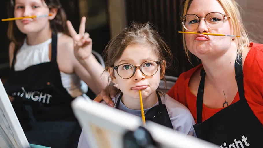 Three girls balancing pencils on their lips indoors.