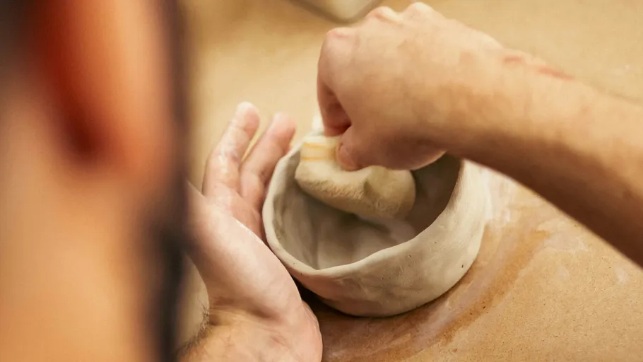 Hands shaping clay bowl on a table.