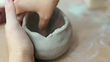 Hands shaping clay bowl on table.
