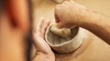 Hands shaping clay bowl with sponge, table surface.
