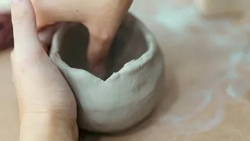 Hands shaping clay bowl on a table.