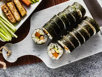 Sushi rolls are placed on a wooden board next to vegetables.