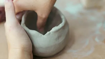 Hands shaping clay bowl on table.