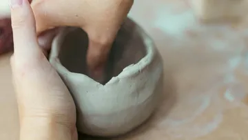 Hands shaping clay bowl, wooden table.
