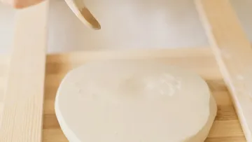 Round bread dough being flattened on wood.