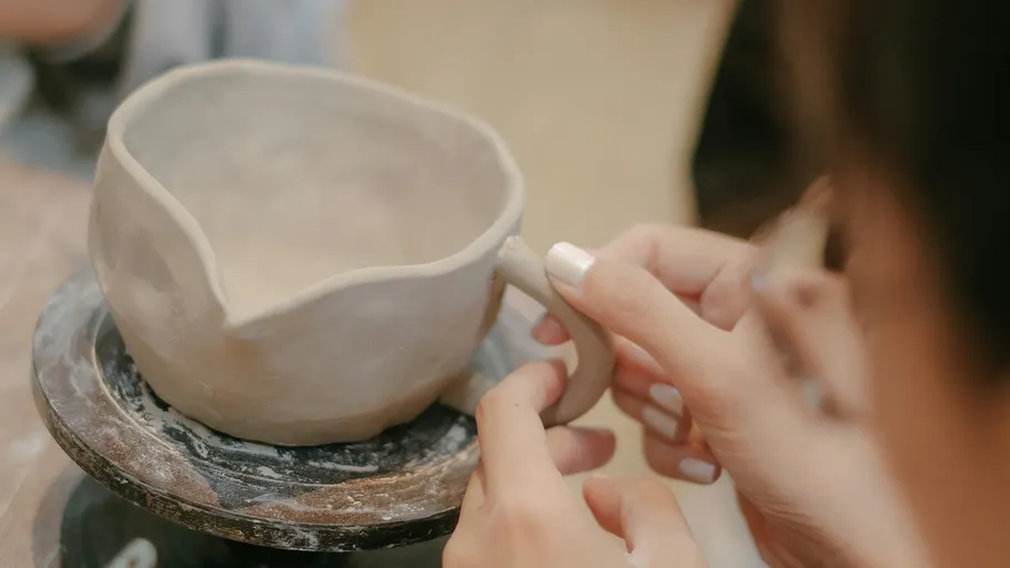 Hands shaping clay handle on pottery cup.