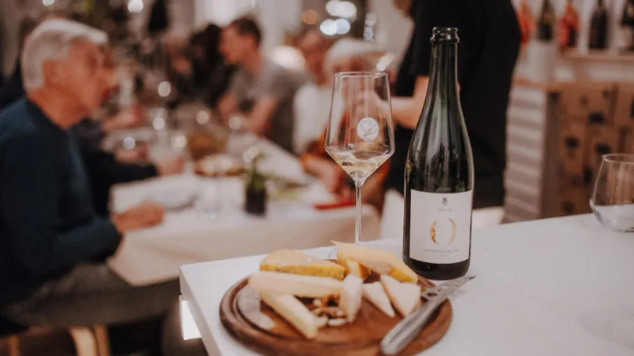 Käse und Wein auf Tisch, Menschen im Hintergrund.