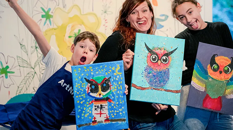 Three people smiling, holding colorful owl paintings.
