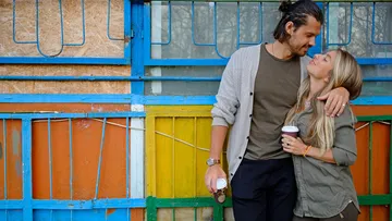 Couple holding coffee cups, colorful wall background.