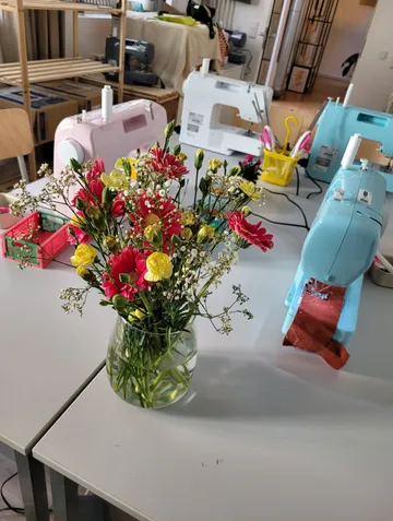 Blumenstrauß auf Tisch mit Nähmaschinen hinter.