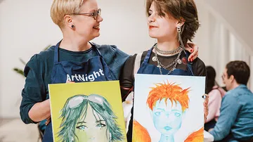 Zwei Personen halten bunte Gemälde drinnen.