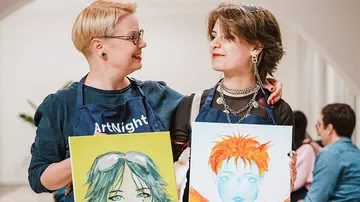 Two people holding paintings, smiling indoors.