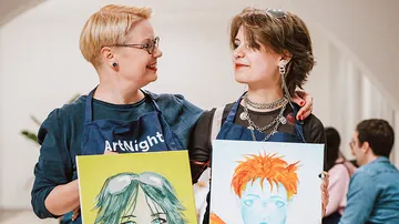 Two people holding paintings, smiling indoors.