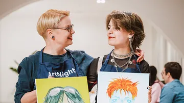 Two people smiling, holding paintings indoors.