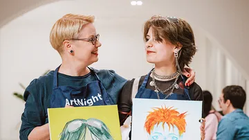 Two people smiling, holding colorful paintings.