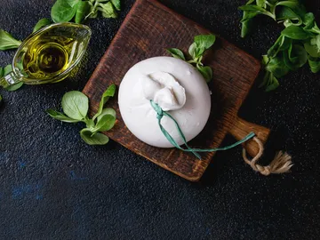 Burrata-Käse liegt auf Holzbrett, umgeben von Kräutern.