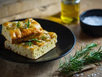 Focaccia-Brot liegt auf schwarzem Teller, umgeben von Kräutern.