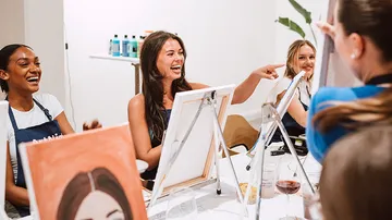 Women laughing, painting at table in studio.