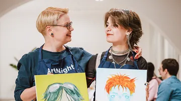 Two people holding paintings, smiling indoors.