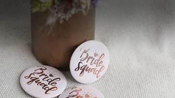 Three bride squad badges beside a vase.