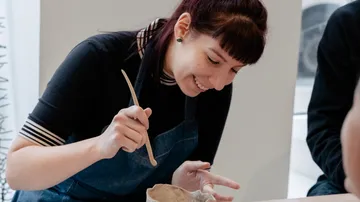Woman sculpting clay at a table, smiling indoors.