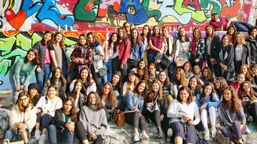 Large group of women posing beside graffiti wall.
