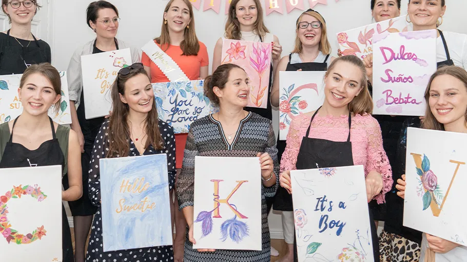Women holding decorated canvases, smiling indoors.