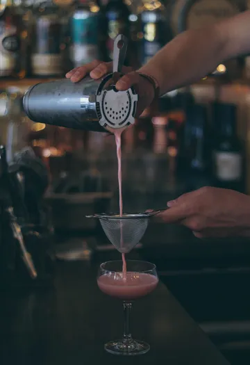 Bartender straining pink cocktail into glass.