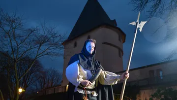 Man in medieval costume holding spear, tower background.