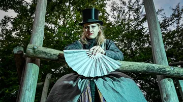 Person in top hat holding white fan outdoors.