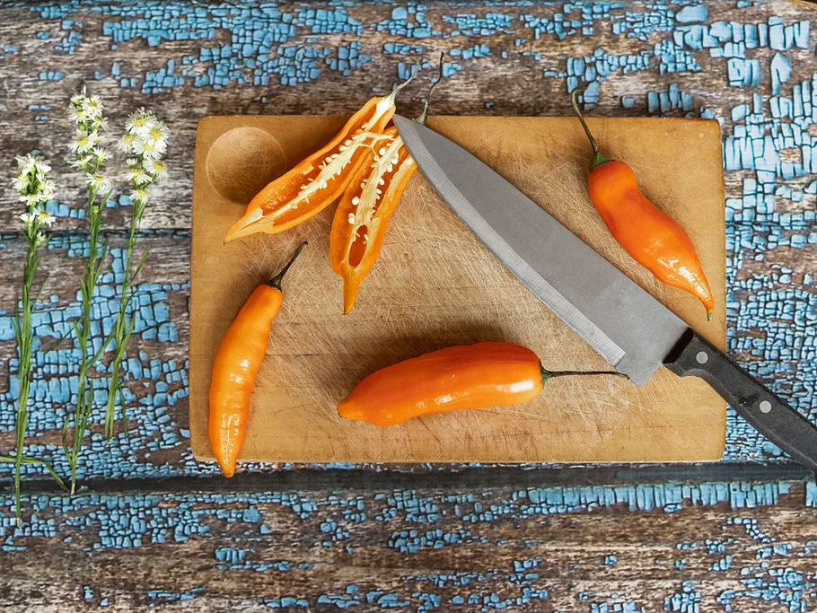 Chilies lie on a wooden board while a knife cuts.
