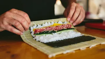 Hände rollen Sushi auf einer Bambusmatte.