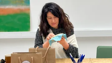 Woman embroidering at table with bags nearby.